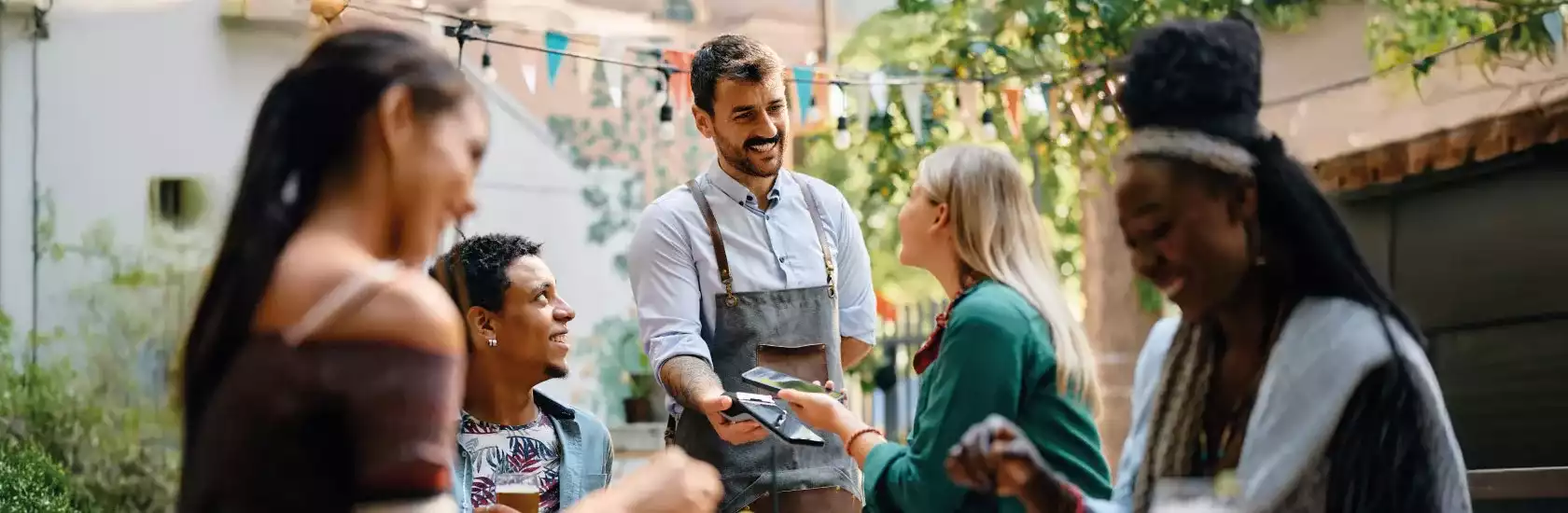 Mesero feliz que recibe el pago sin contacto del teléfono inteligente de una mujer que está siendo con sus amigos en un café.